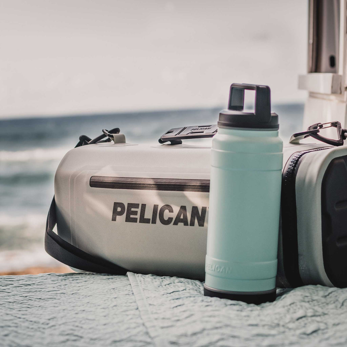 SOFT-CSLING-LGRY Pelican™ Dayventure Sling Soft Cooler on the beach with a 32oz Pelican Bottle