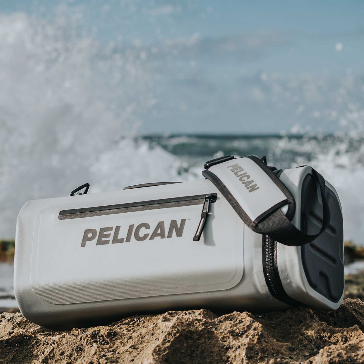 SOFT-CSLING-LGRY Pelican™ Dayventure Sling Soft Cooler on the beach with waves crashing behind it